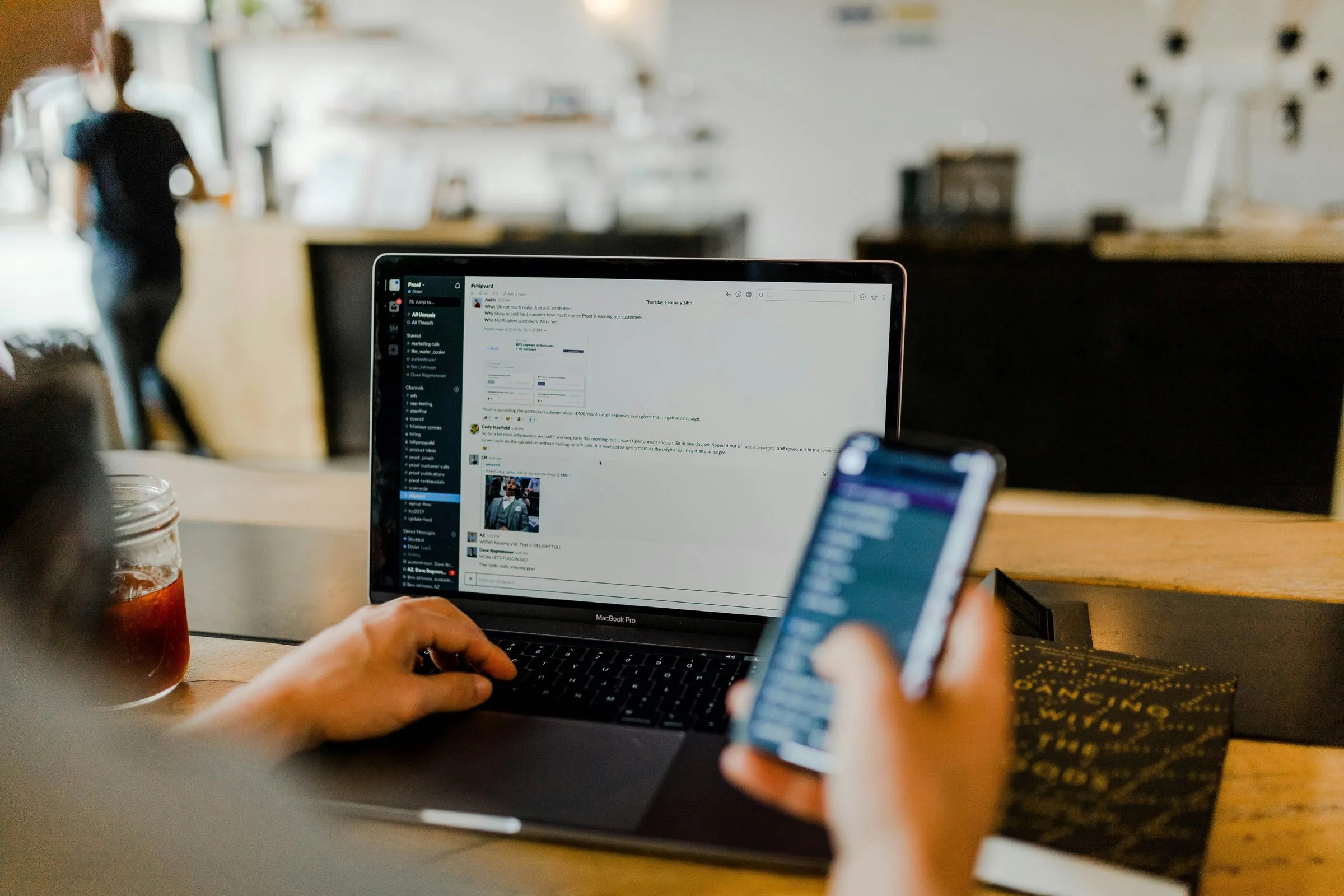 A person working at a laptop in a dim room, holding a phone in one hand.